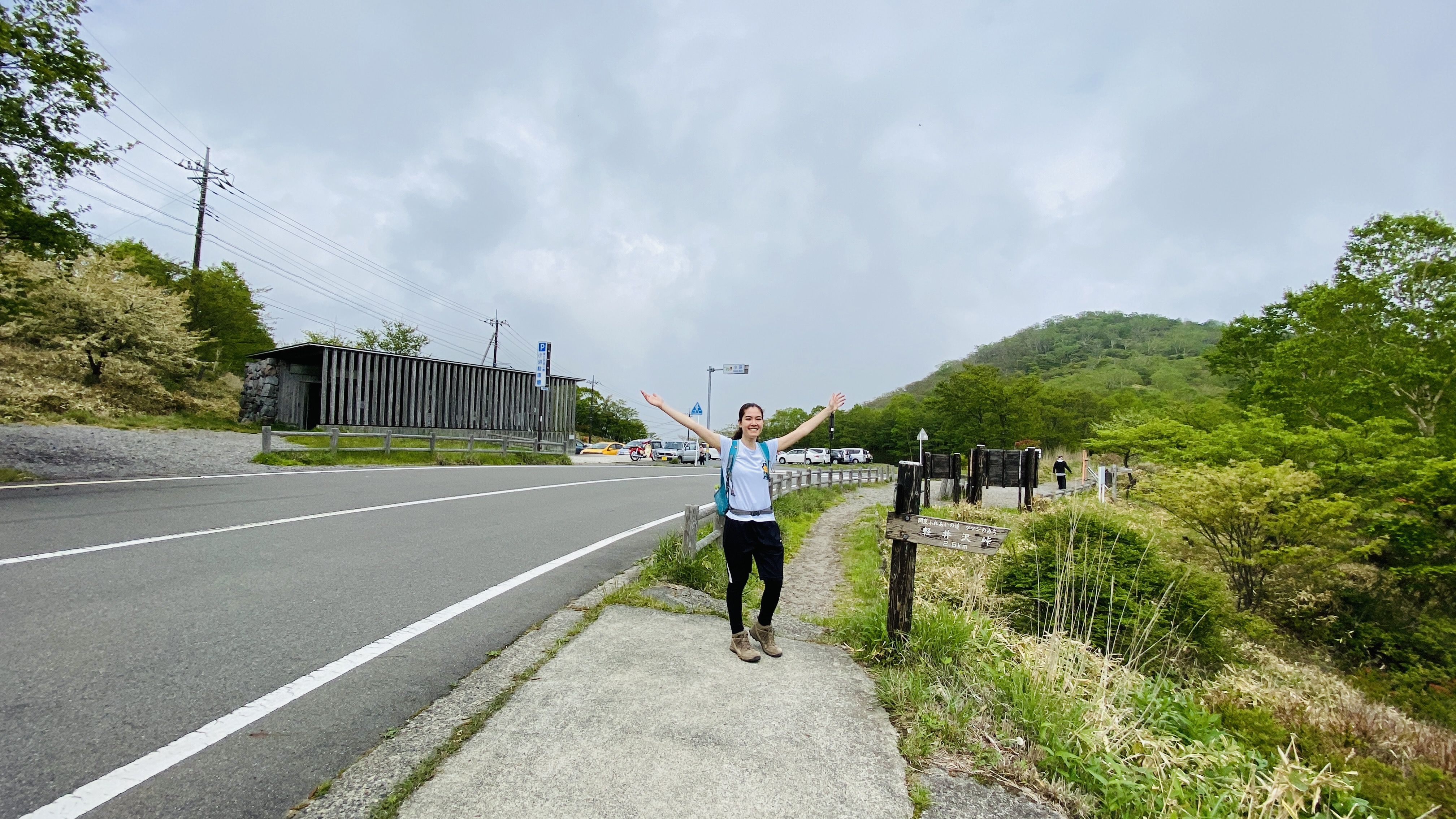 A hiker raises her arms in triumph with Lake Konuma parking lot and bathroom in background