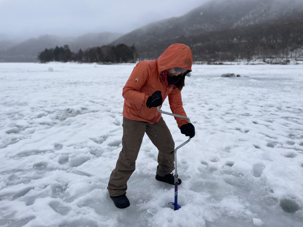 A person in an orange jacket drilling a hole into ice on a lake